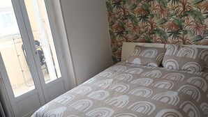 Ein Schlafzimmer mit Schachbrettmuster auf dem Boden, einem Bett mit gemustertem Überwurf und einer Tür mit Blick ins Freie, sowie einer Wand mit tropischem Blattmuster.