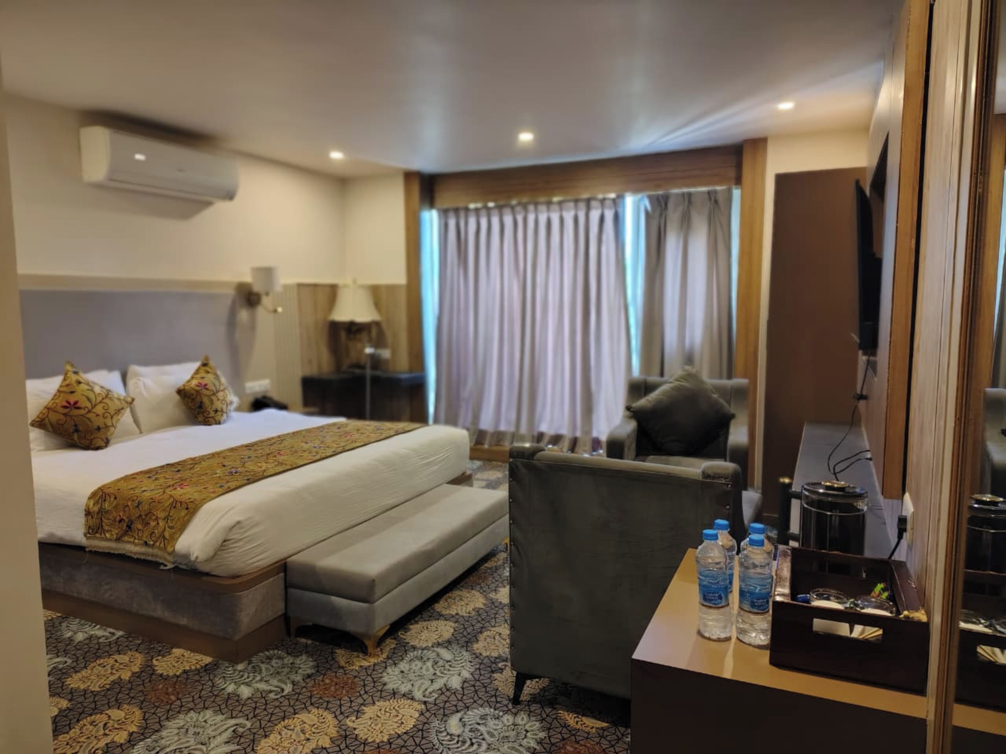 A hotel room with a bed, a desk with water bottles, a chair, a TV, and a window with curtains.