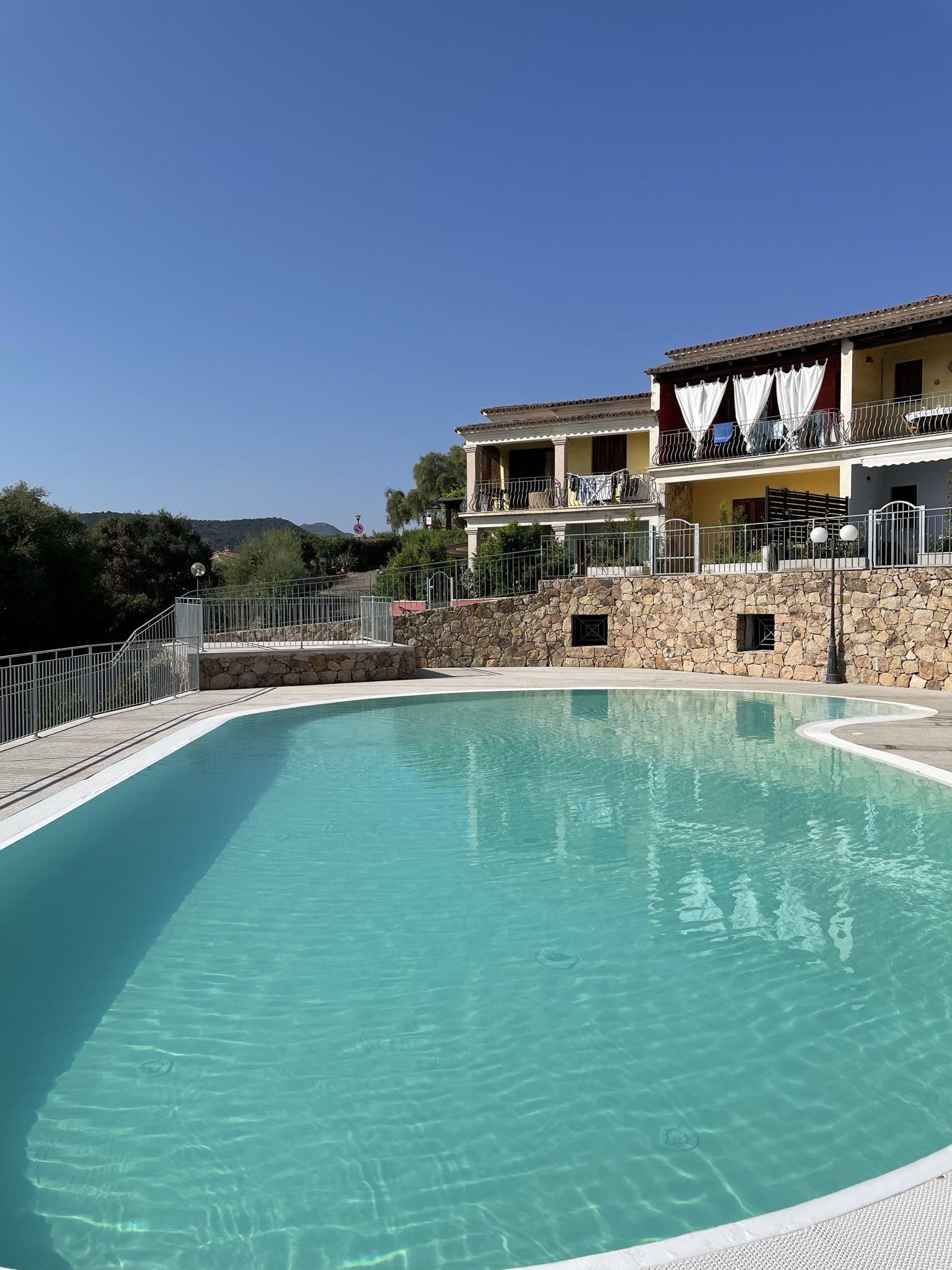A clear swimming pool with a stone wall and a building with balconies.