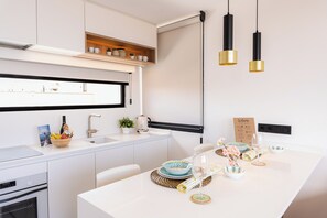 Une cuisine moderne avec des meubles blancs, un four encastré, un évier et un bar pour prendre le petit-déjeuner. On y trouve des suspensions lumineuses, un panneau d’accueil et une petite plante en pot sur le comptoir.