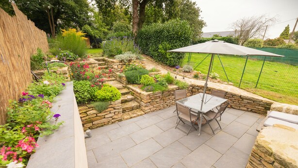 Een terras met een tafel en stoelen, omgeven door stenen muren en diverse beplanting.
