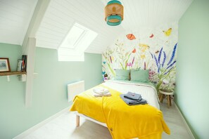 A bedroom with a sloped ceiling, a skylight, a bed with a yellow bedspread, a radiator, and a wall with floral wallpaper. - Gîte Youna - stone charm & secret garden (Lizio)