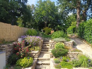 A stone pathway leading through a garden with various plants and a wooden fence on one side. - Gîte Youna - stone charm & secret garden (Lizio)