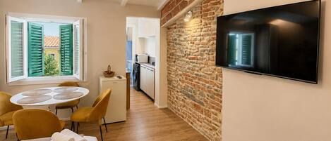 A compact hotel room with a dining area, a TV mounted on the wall, a small kitchen, and a view through a window with shutters.