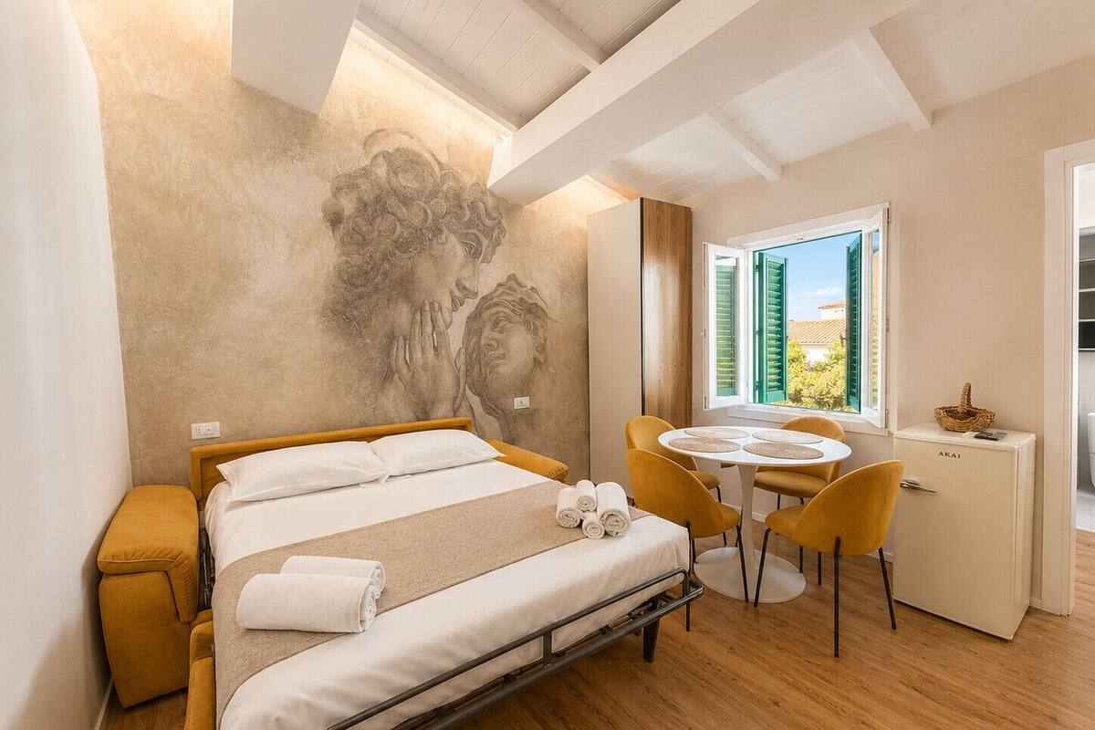 A bedroom with a bed, a desk, chairs, a window with shutters, and a wall mural.