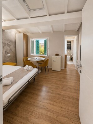 A hotel room with a bed, a small table with chairs, a window with shutters, a bathroom visible through an open door, and a kitchenette area. - Pitti Suite 1 (Florence)