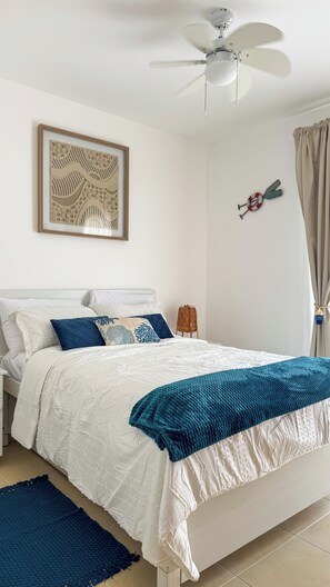 A bedroom with a bed, a ceiling fan, a framed artwork, a small ladder, and a curtain.