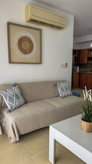 A living room with a beige sofa, two patterned throw pillows, a framed abstract artwork, a white table, a potted plant, and a kitchen area in the background.