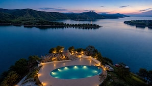 Ein großer, ovaler Swimmingpool mit umlaufender Liegefläche, vor einer ruhigen Seenlandschaft und fernen Bergen.