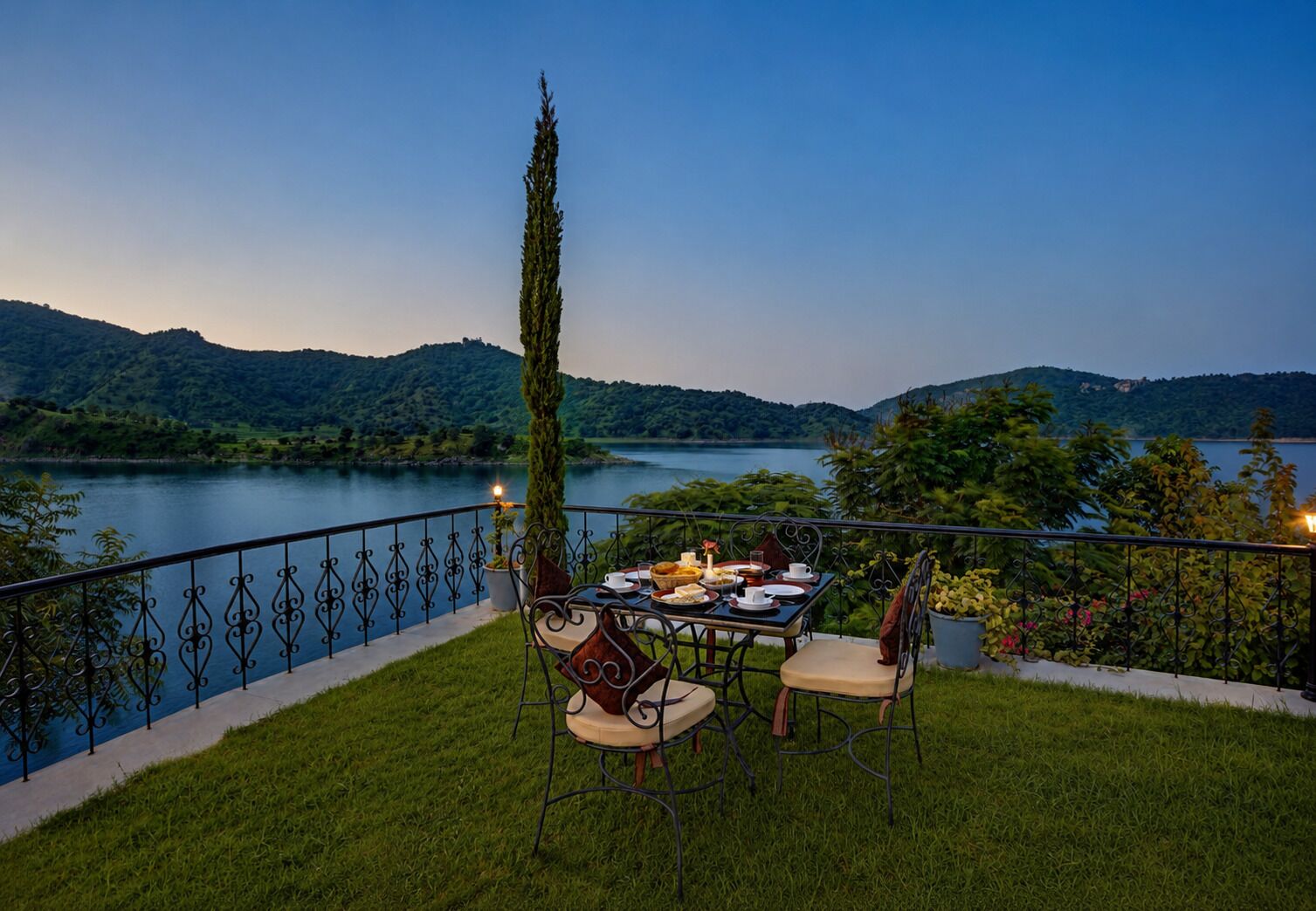 Une table à manger avec des chaises, placée sur un balcon donnant sur un lac, avec une balustrade et un pot de fleurs.