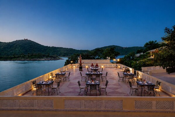 Un área de comedor al aire libre con mesas y sillas, con vista a un cuerpo de agua y colinas.