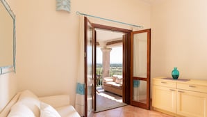 A room with a white sofa, a yellow cabinet, and a glass door leading to an outdoor area with a wicker chair and a view of the landscape. - Casa Luna e Sole by Wonderful Italy (Budoni)