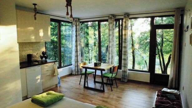 Ein Zimmer mit Esstisch, Stühlen, einer Bank, einer Küchenzeile und großen Fenstern mit Blick auf Bäume.