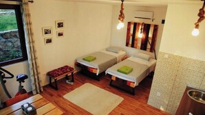 Ein kleines Zimmer mit zwei Einzelbetten, einem kleinen Tisch, einer Bank, einem Fenster mit Vorhängen und einer Küchenzeile.