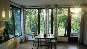 Ein Zimmer mit Esstisch, Stühlen, einem Fenster mit Vorhängen und Blick auf Bäume.