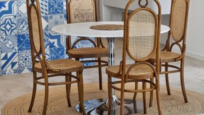 Un comedor con una mesa redonda, cuatro sillas de mimbre, una lámpara colgante de fibra natural y una cocina al fondo.