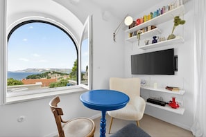 Una habitación con una mesa redonda azul, una silla blanca, una estantería y un televisor en la pared.