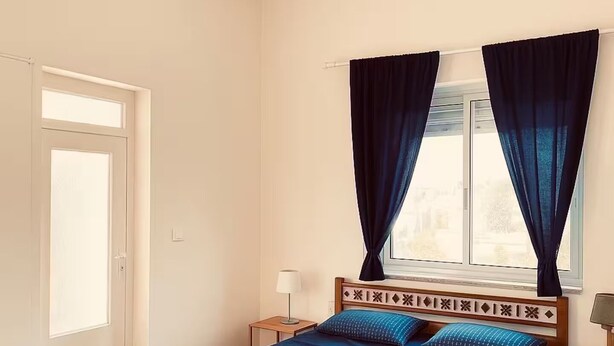 Ein Schlafzimmer mit hölzernem Bettgestell, einem Fenster mit Vorhängen, einem Nachttisch und einem Beistelltisch.