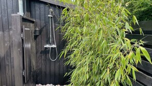 Ducha exterior de madera con manguera, rodeada de guijarros y una planta de bambú.