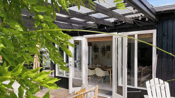 Terraza de madera con puerta de vidrio que da a un comedor interior con sillas blancas y una mesa.