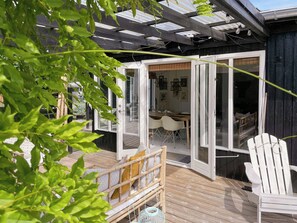 Une terrasse en bois avec une porte vitrée donnant sur un espace de repas intérieur, avec des chaises blanches et une table.
