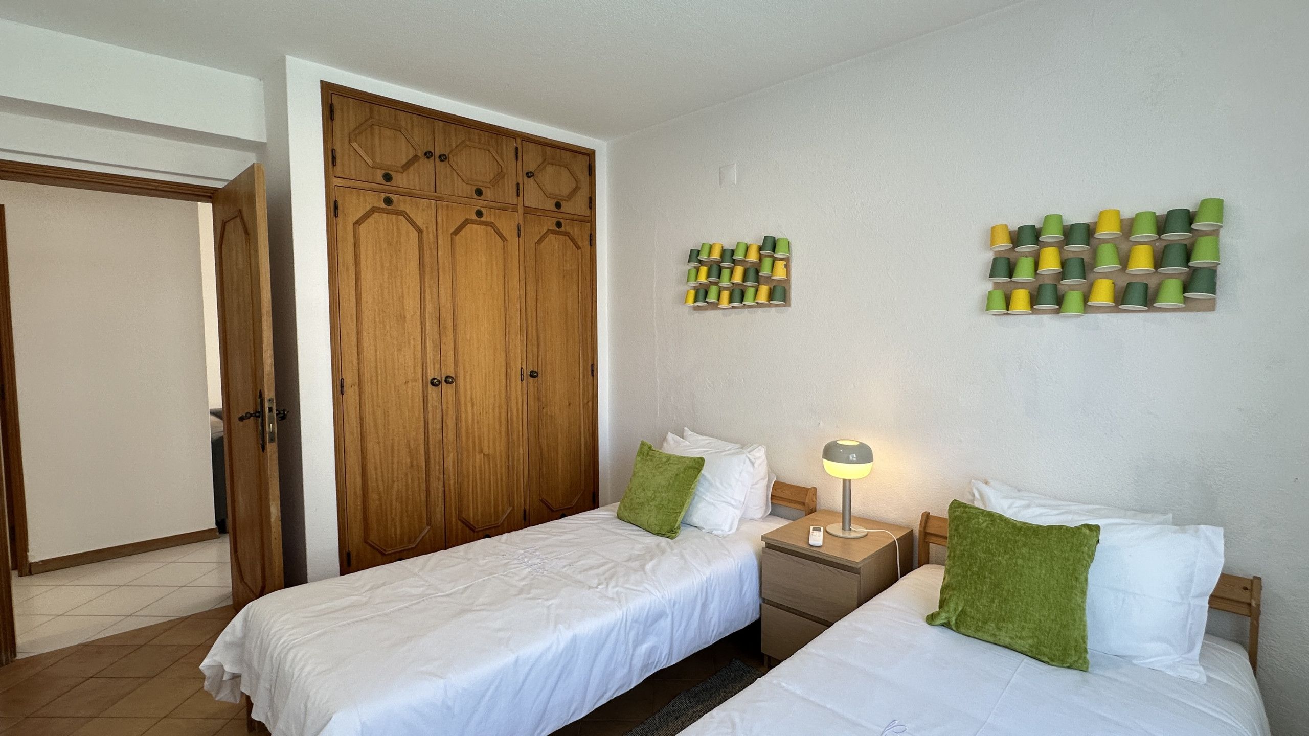 A bedroom with two beds, a wooden wardrobe, a bedside table with a lamp, and a wall-mounted spool holder.