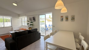A modern living room with a sofa, a dining table, and chairs. There is a large window with a view of the outside, and a plant on the windowsill. - Rosa | Private Pool | Sleeps 8 | Albufeira (Albufeira)