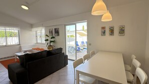 A modern living room with a sofa, a dining table, and chairs. There is a large window with a view of the outside, and a plant on the windowsill. - Rosa | Private Pool | Sleeps 8 | Albufeira (Albufeira)