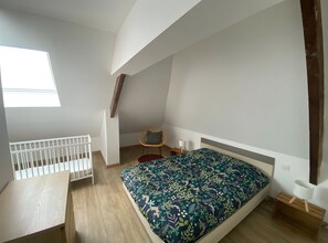 A small, well-lit room with a sloped ceiling, a single bed with a floral bedspread, a small table, a chair, and a window providing natural light.