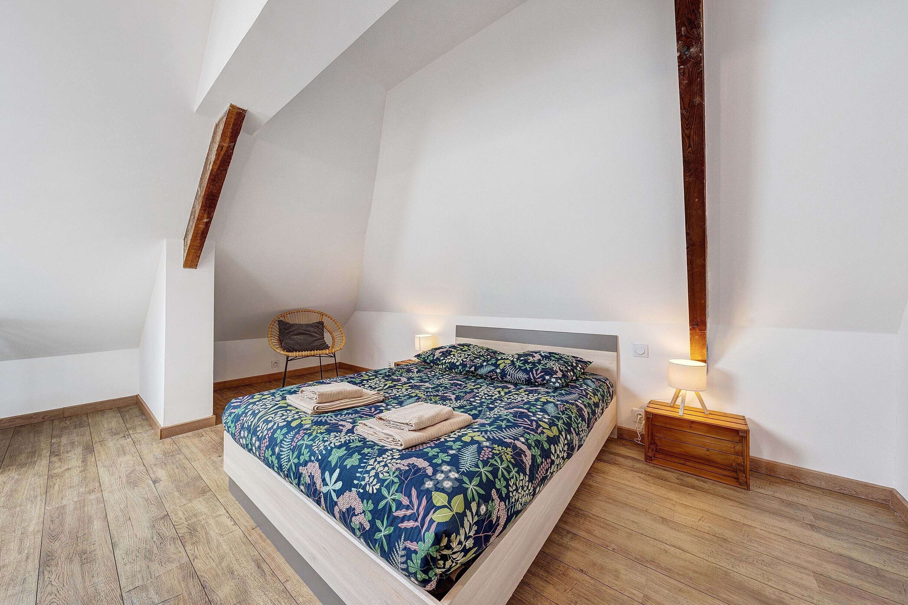 Een slaapkamer met een schuine kap, een bed met bloemenbedekking, een nachtkastje met een lamp en een stoel.