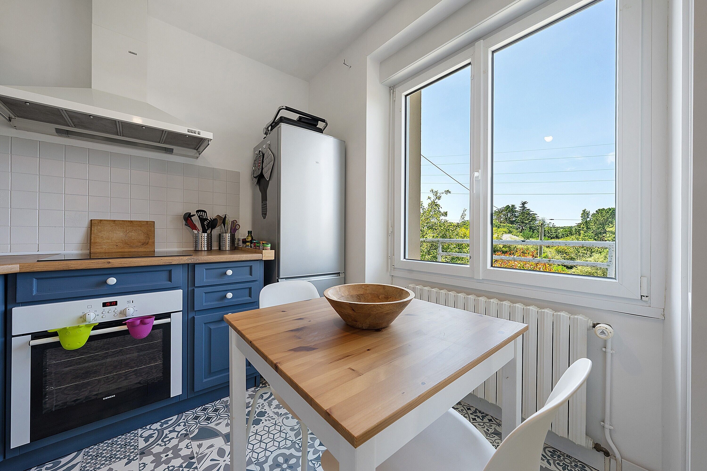 Une cuisine avec une table en bois, un saladier, un réfrigérateur et une fenêtre donnant sur un beau paysage.