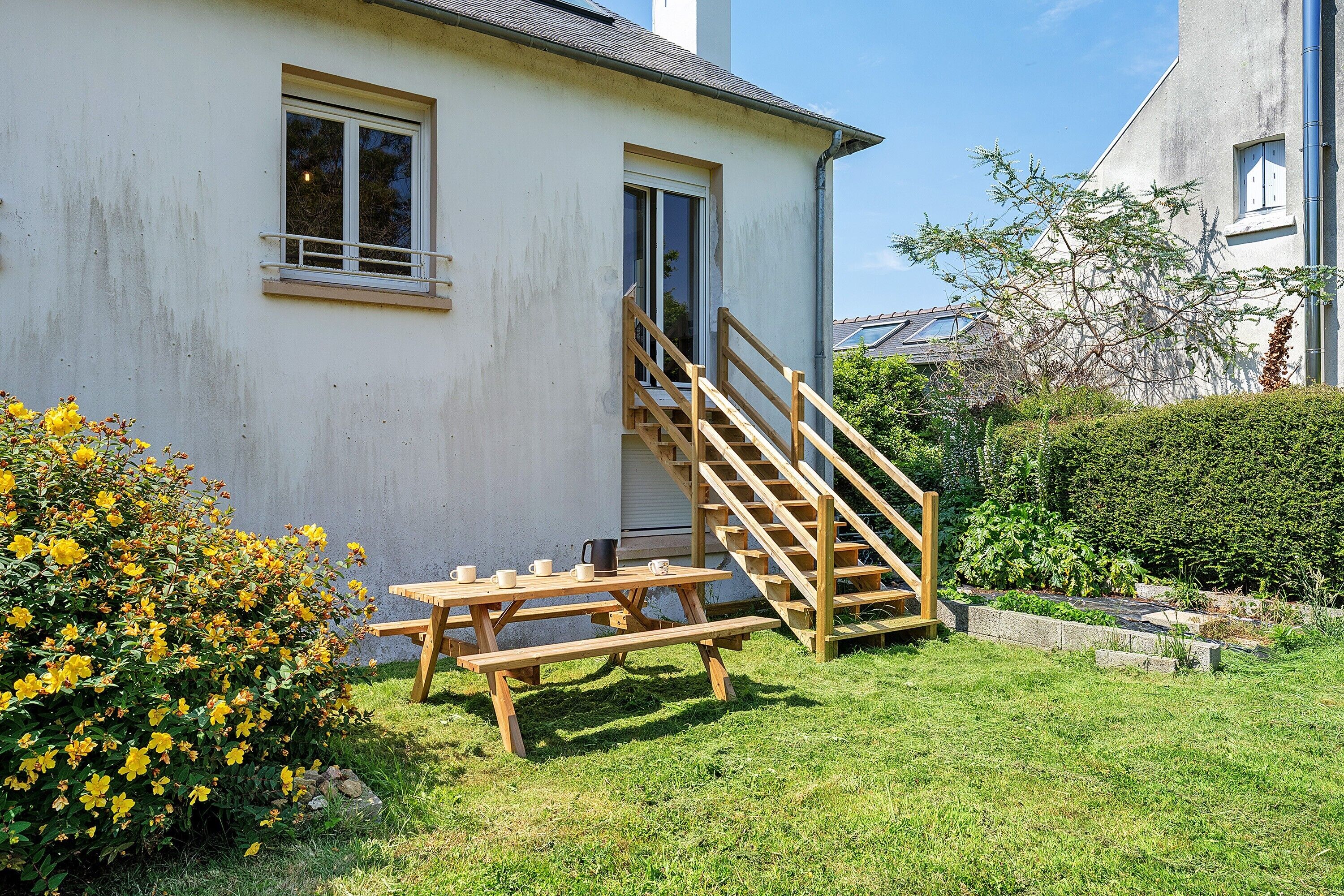 Une maison avec un escalier en bois menant à une porte, une table de pique-nique et un jardin parsemé de fleurs jaunes.
