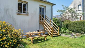Une maison avec un escalier en bois menant à une porte, une table de pique-nique et un jardin parsemé de fleurs jaunes.