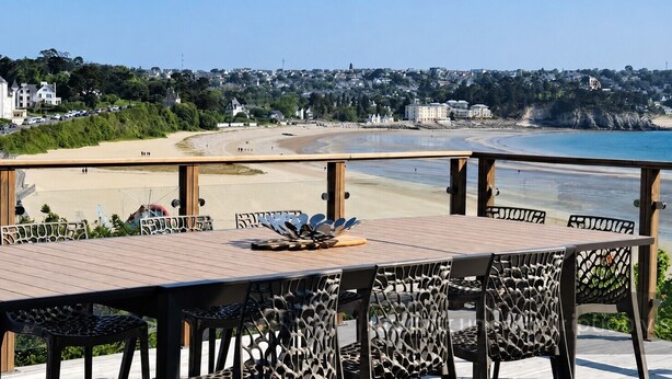 Eine Holzterrasse mit Tisch und Stühlen mit Blick auf einen Strand und eine Küstenstadt.