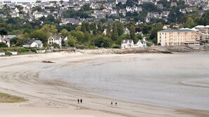 Ein Strand mit einigen Personen, die am Ufer entlanggehen, feinem Sand und Häusern im Hintergrund.