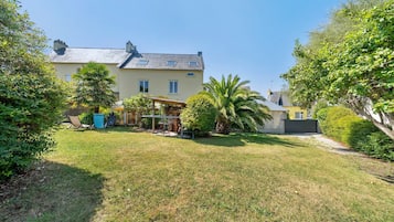 Uma casa de dois andares com varanda coberta, gramado e diversas árvores e arbustos.
