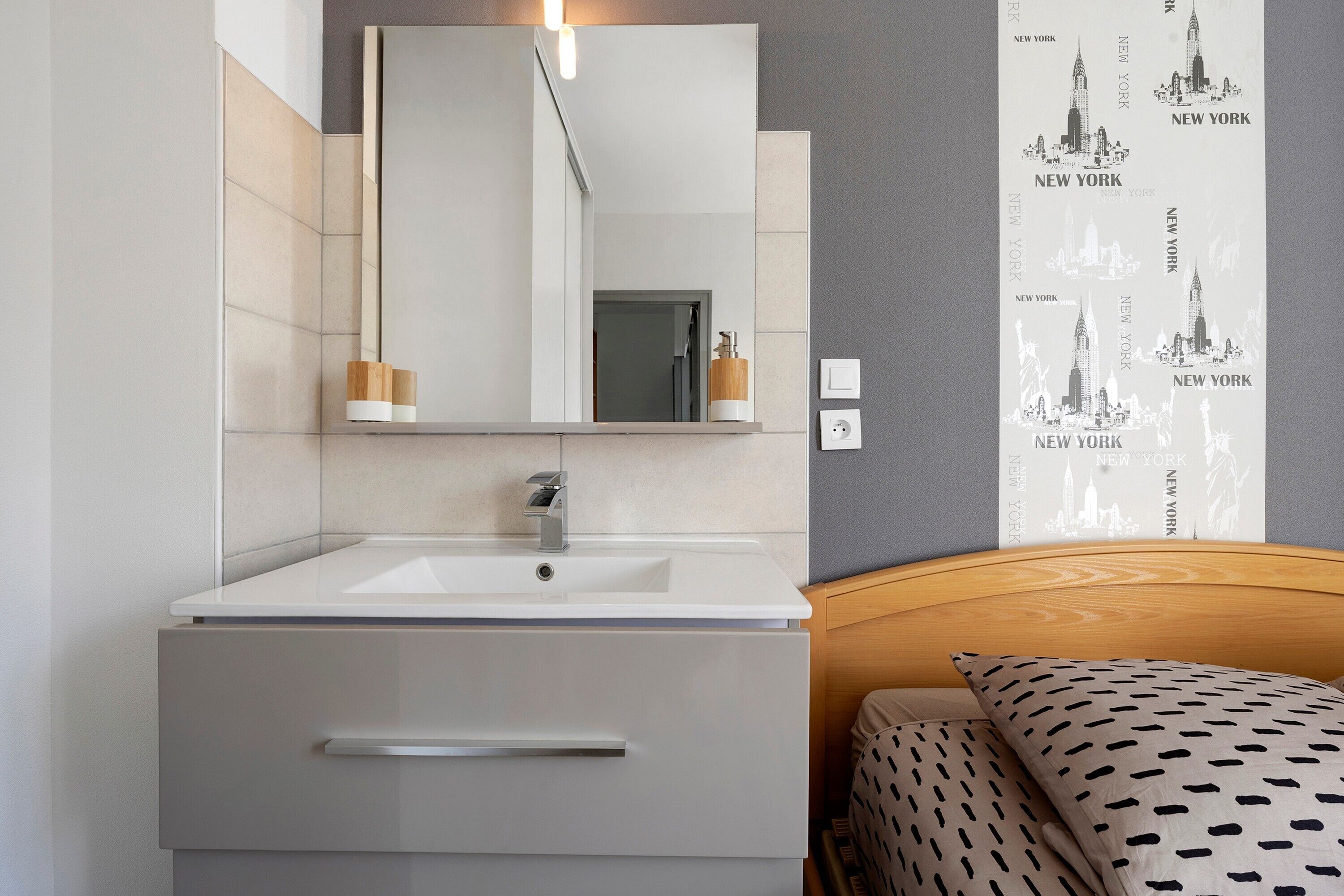 A modern bathroom with a white sink, a mirror, and a wooden bedside table.