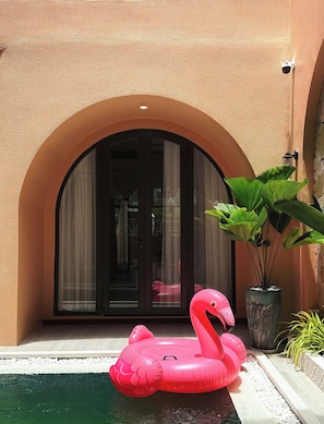 Una piscina con un flotador de flamenco inflable rosa, una planta grande en maceta y la entrada de un edificio con un arco.