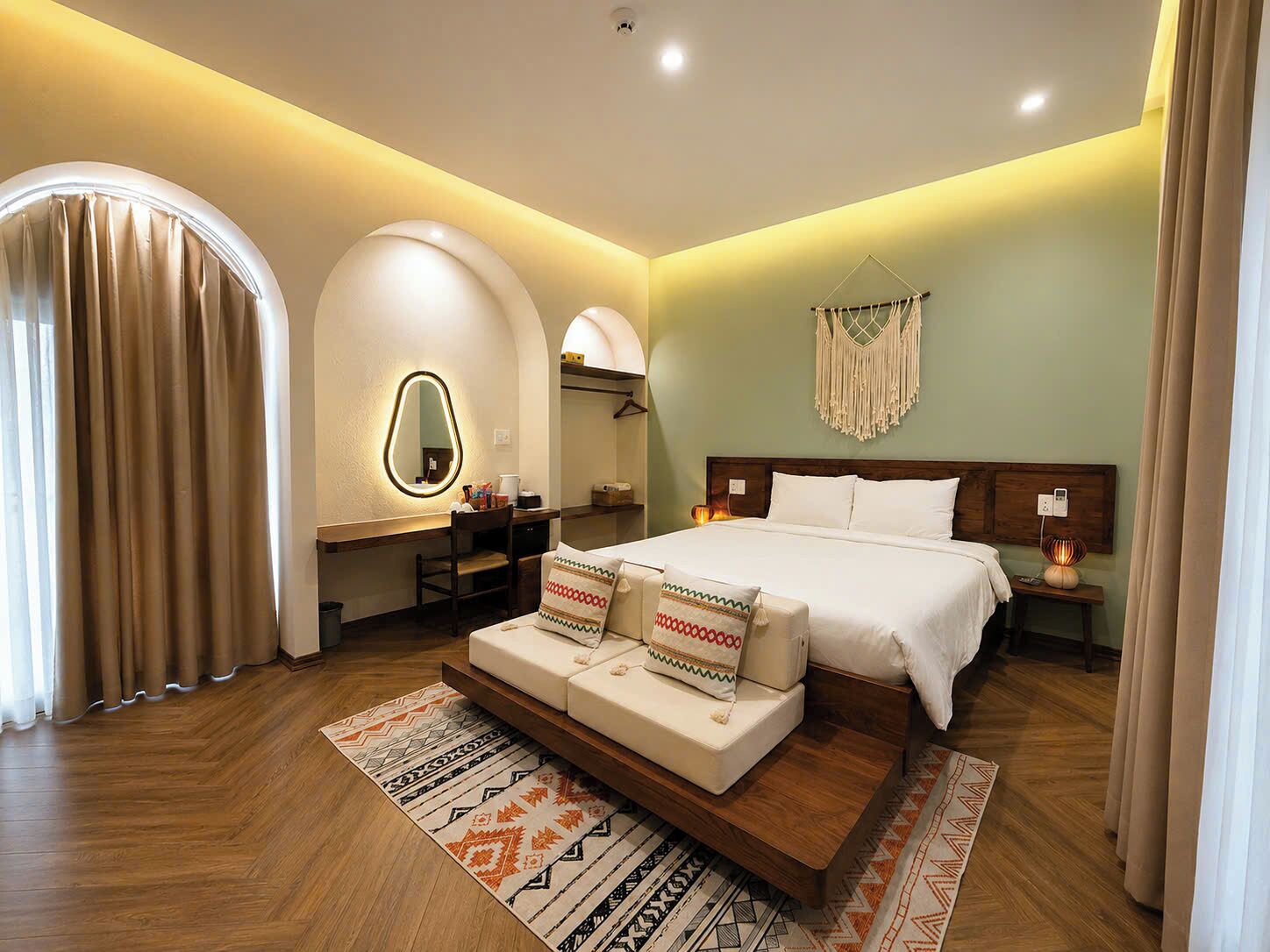 A modern bedroom with a large bed, a vanity with a mirror, a desk, and a macrame wall hanging.