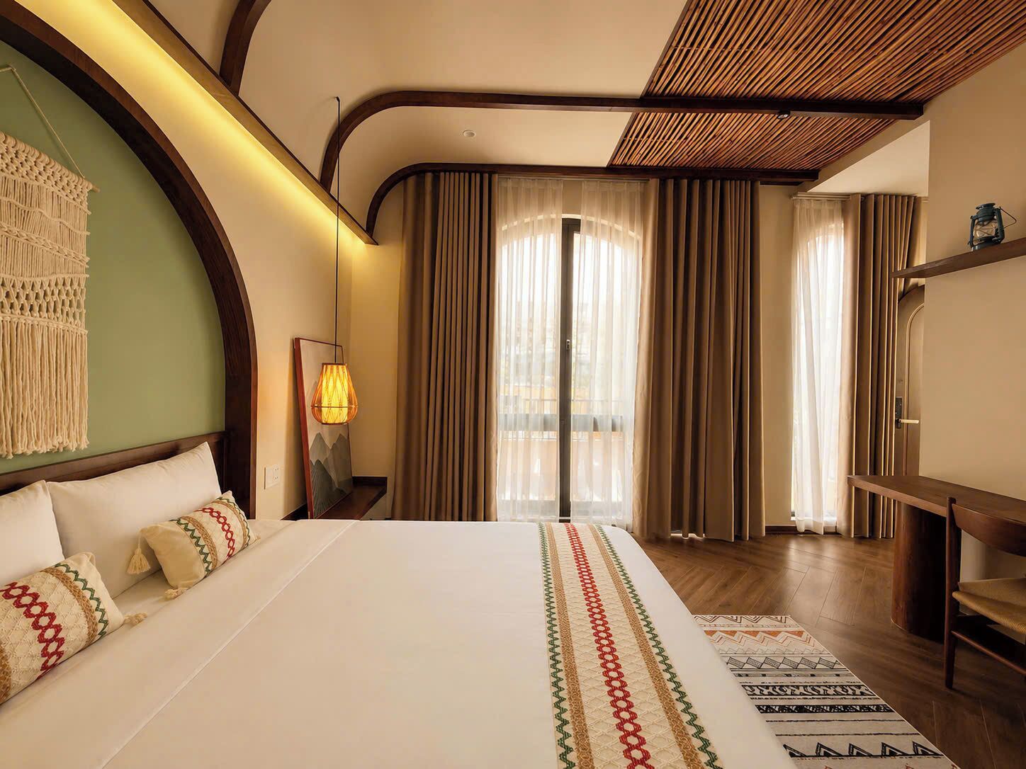A hotel room with a bed, a macrame wall hanging, a lamp, and a view of the outside through large curtains.