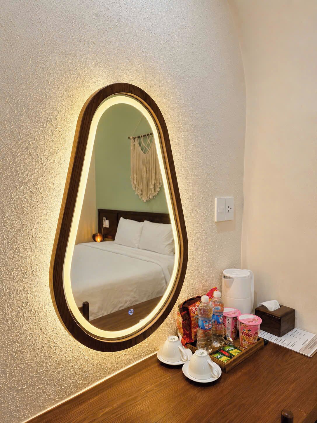 Habitación de hotel con un espejo grande, una cama, una mesita de noche con artículos de aseo y una lámpara fijada a la pared.