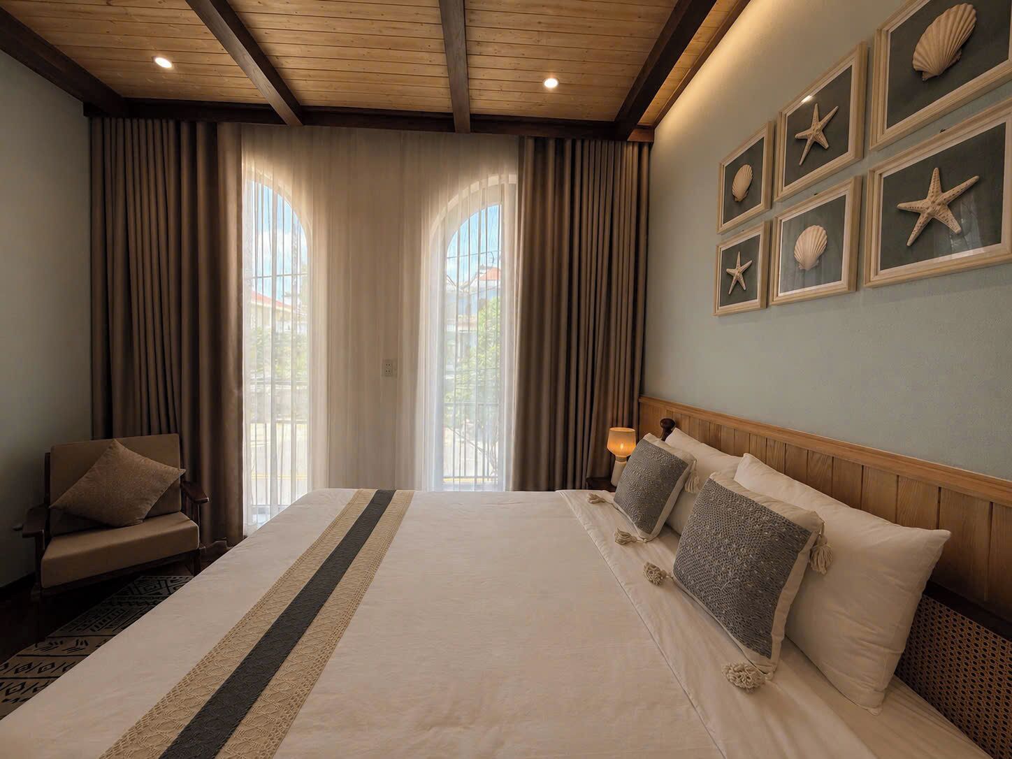 Habitación de hotel con una cama grande, una silla, una mesita de noche y una pared decorada con estrellas de mar y conchas enmarcadas.