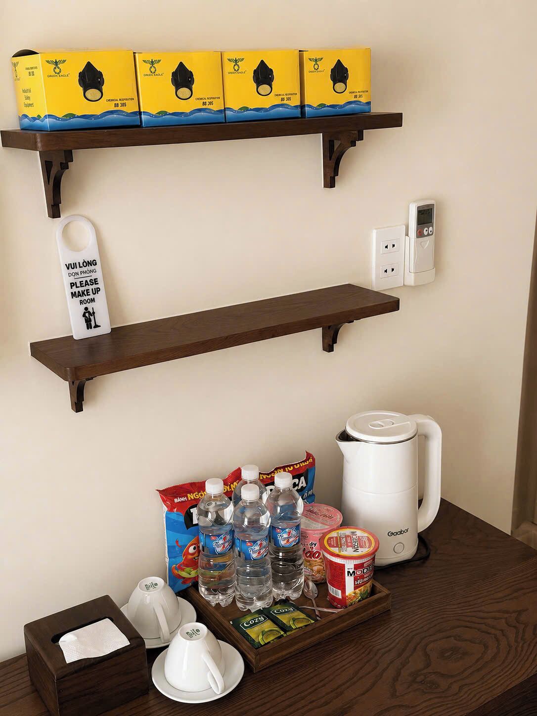 A hotel room with a shelf holding boxes, a sign, a bedside table with a kettle, cups, bottles, and a tissue box.