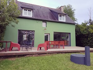 Uma casa verde com telhado de telhas, porta da frente e uma grande porta de vidro deslizante. Há cadeiras e uma mesa no alpendre.