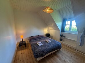 Un dormitorio con techo inclinado, una cama con colcha estampada, una lámpara de noche, una mesita de noche pequeña, una ventana con cortinas y un colgador de luz.