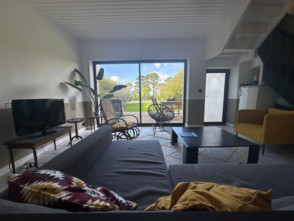 Sala de estar com sofá, televisão, mesa de centro e uma cadeira. Porta de vidro deslizante dá acesso a área externa com gramado e mesa.