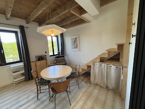 Un comedor con una mesa redonda, sillas, una lámpara colgante, un ventanal con cortinas y una escalera de madera.