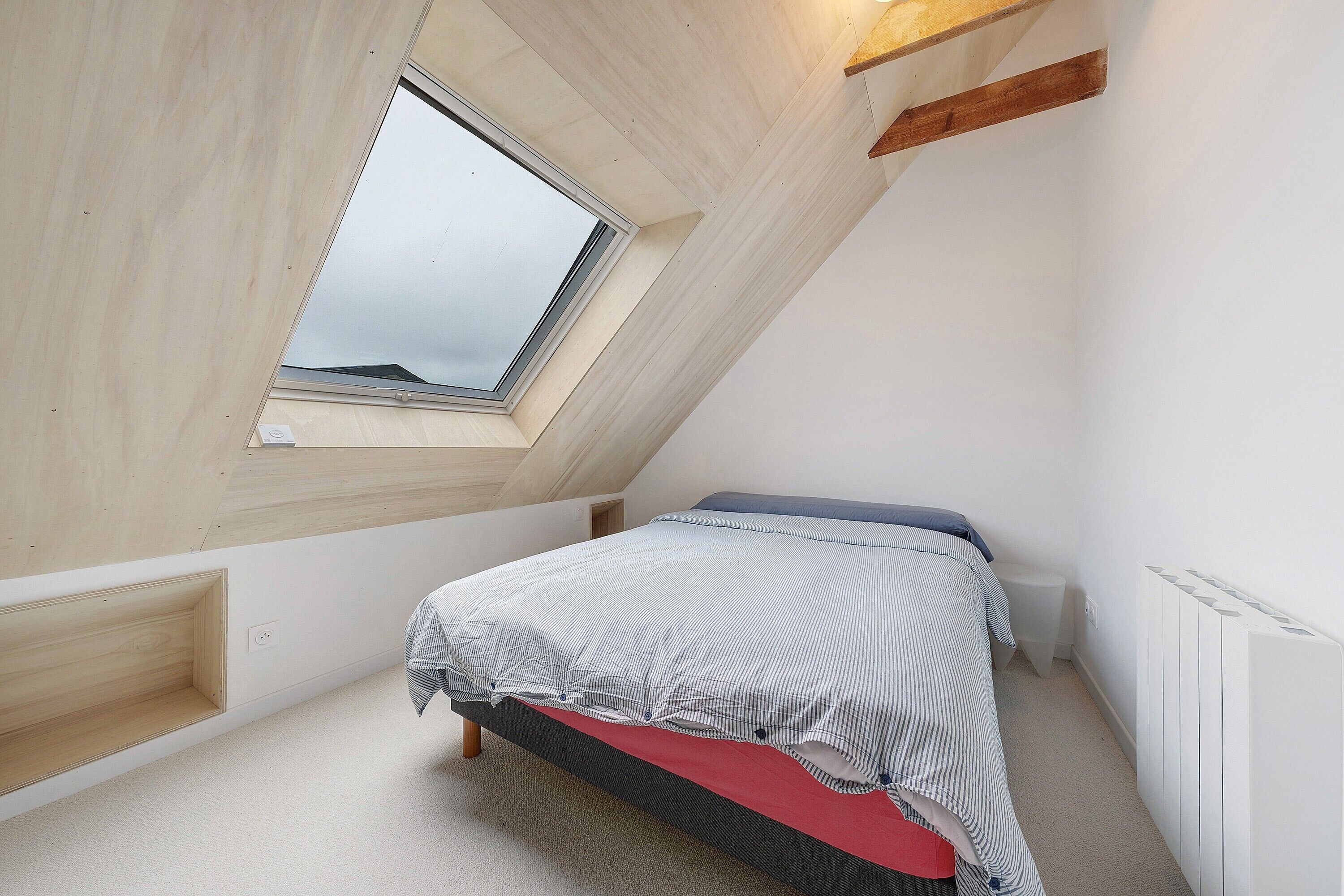 Een slaapkamer met een schuine kap, dakraam, een bed, een radiator en een klein nachtkastje.