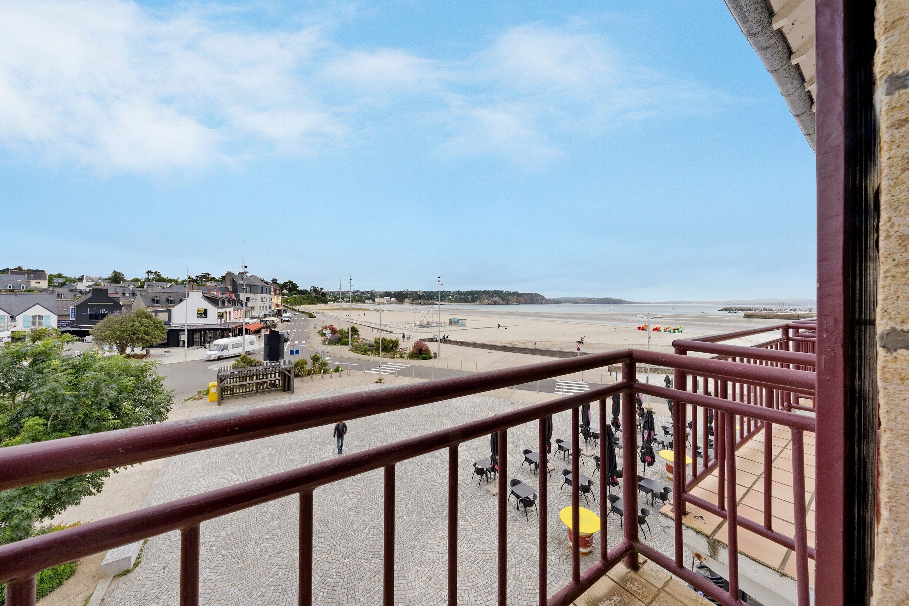A balcony with a view of a beach, a paved area with tables and chairs, and buildings in the background.