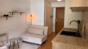 Un espacio habitable pequeño con kitchenette, un sofá, una mesita redonda y una puerta que da a otra habitación.